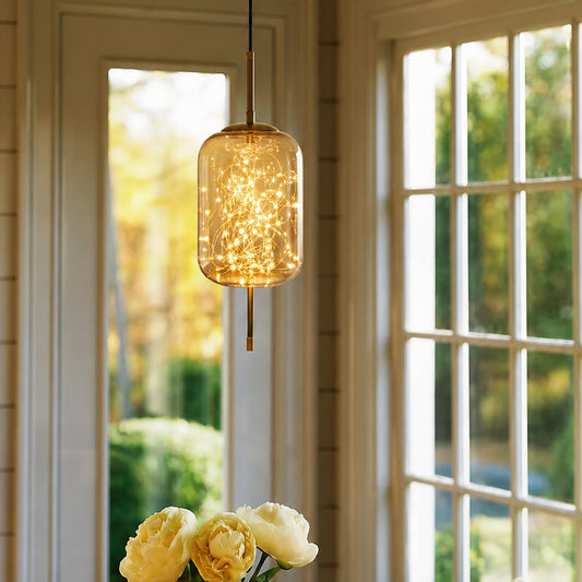 A hanging pendant light with a clear glass cylinder and warm glowing fairy lights inside, suspended in a bright room with white-framed windows and blurred yellow flowers below.