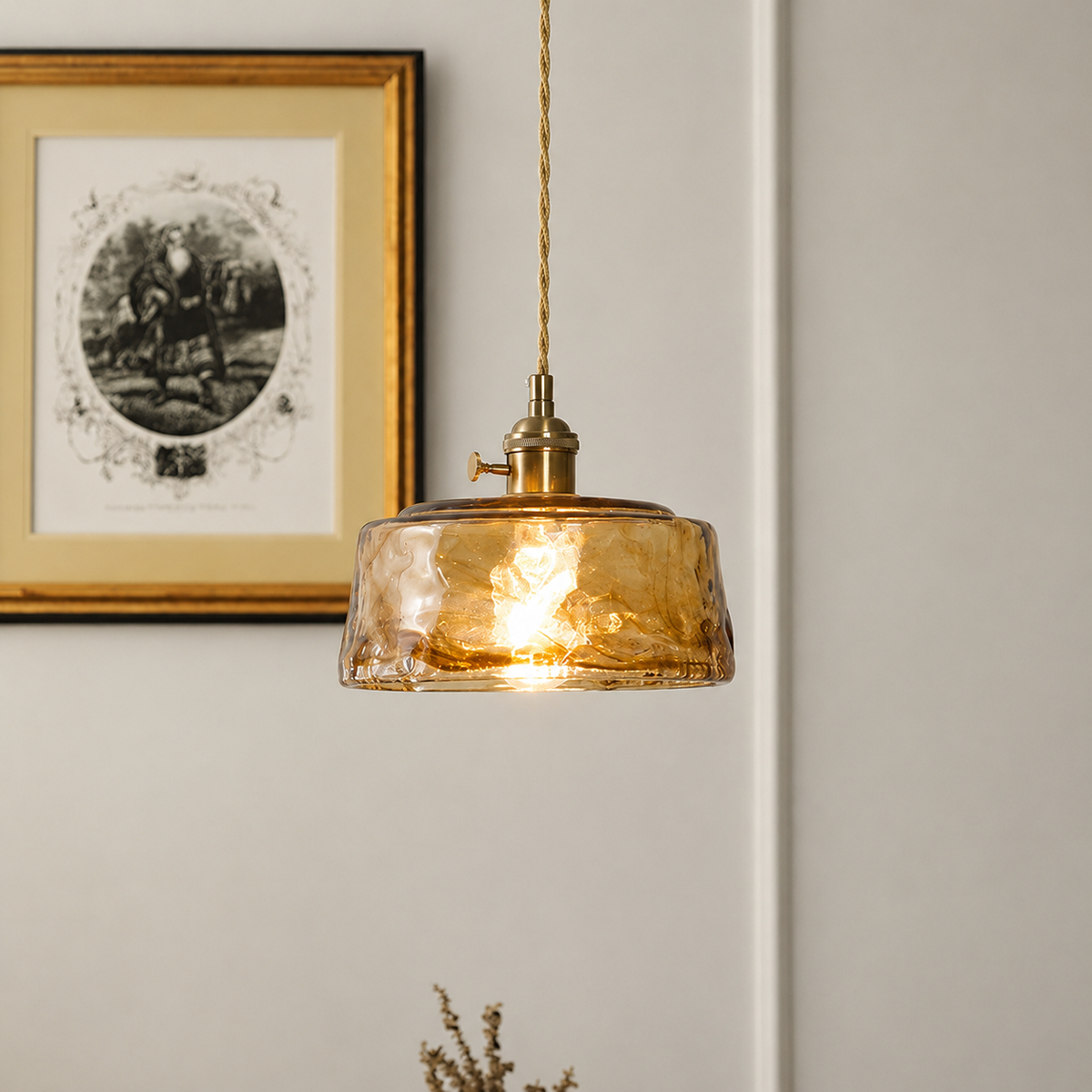 A hanging pendant light with a textured amber glass shade and brass-toned metal fixture, suspended by a cord against a light neutral wall with framed wall art visible in the background.