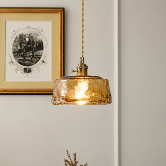 A hanging pendant light with a textured amber glass shade and brass-toned metal fixture, suspended by a cord against a light neutral wall with framed wall art visible in the background.