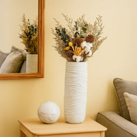 White textured ceramic vase styled with dried flowers placed on a wooden table beside a sofa in a cozy beige living room setting.