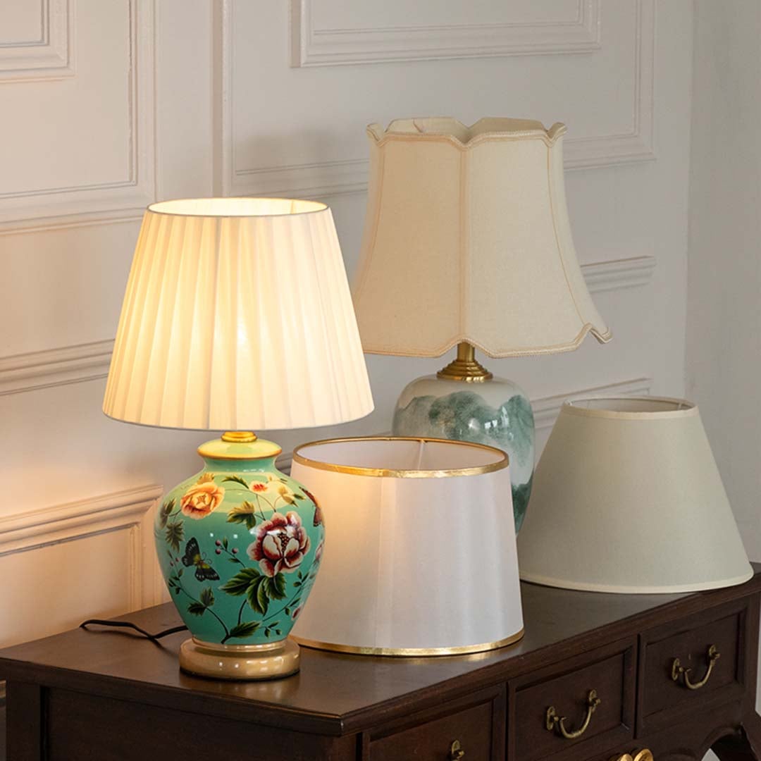 Green floral ceramic table lamp glowing with warm light styled on wooden console beside elegant lampshades.