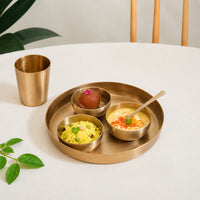 A brass thali set displayed with naan curry rice raita and a brass tumbler on a wooden table creating a warm lifestyle dining setup with an authentic feel.