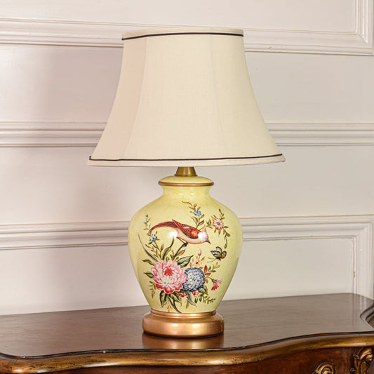 Decorative yellow ceramic table lamp with floral bird design gold metal base and fabric shade on off white background.