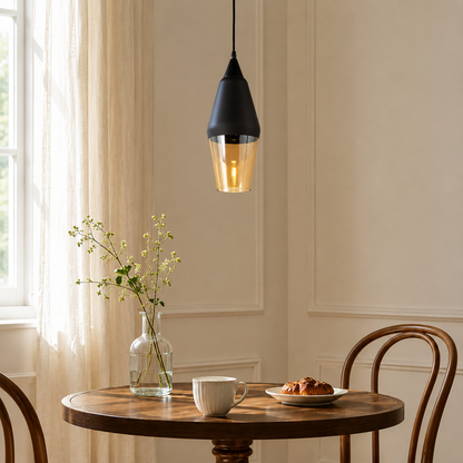 A modern black pendant light with a warm glowing glass shade hangs over a round wooden dining table in a softly lit room with cream walls, a window, sheer curtains, a glass vase with flowers, a white mug, and a croissant on a plate.