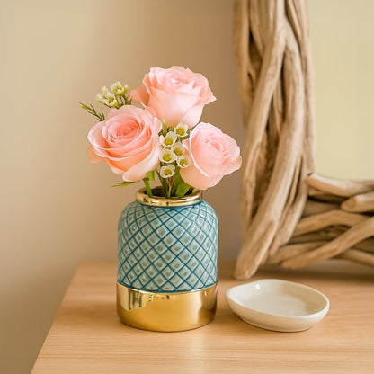 Gold and blue ceramic vase with a diamond pattern holding pink roses and white buds placed on a wooden table near a rustic mirror.