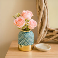 Gold and blue ceramic vase with a diamond pattern holding pink roses and white buds placed on a wooden table near a rustic mirror.