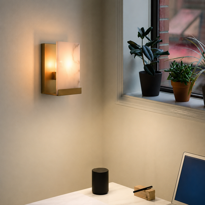 A modern wall-mounted sconce with a rectangular gold-toned metal frame and a glowing warm white marble-look glass shade, installed on a beige wall in a room setting next to a window with potted plants.