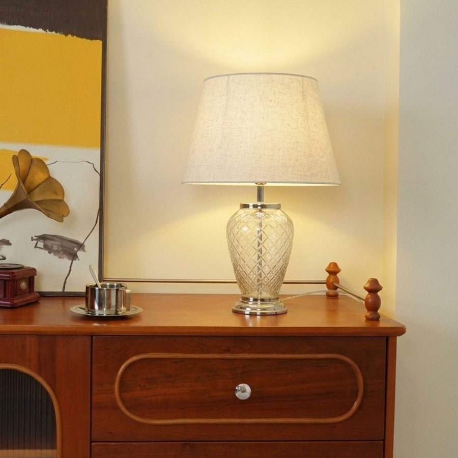 A table lamp with a textured cream fabric drum shade and a carved crystal or glass base sits on a wooden sideboard against a warm beige wall.