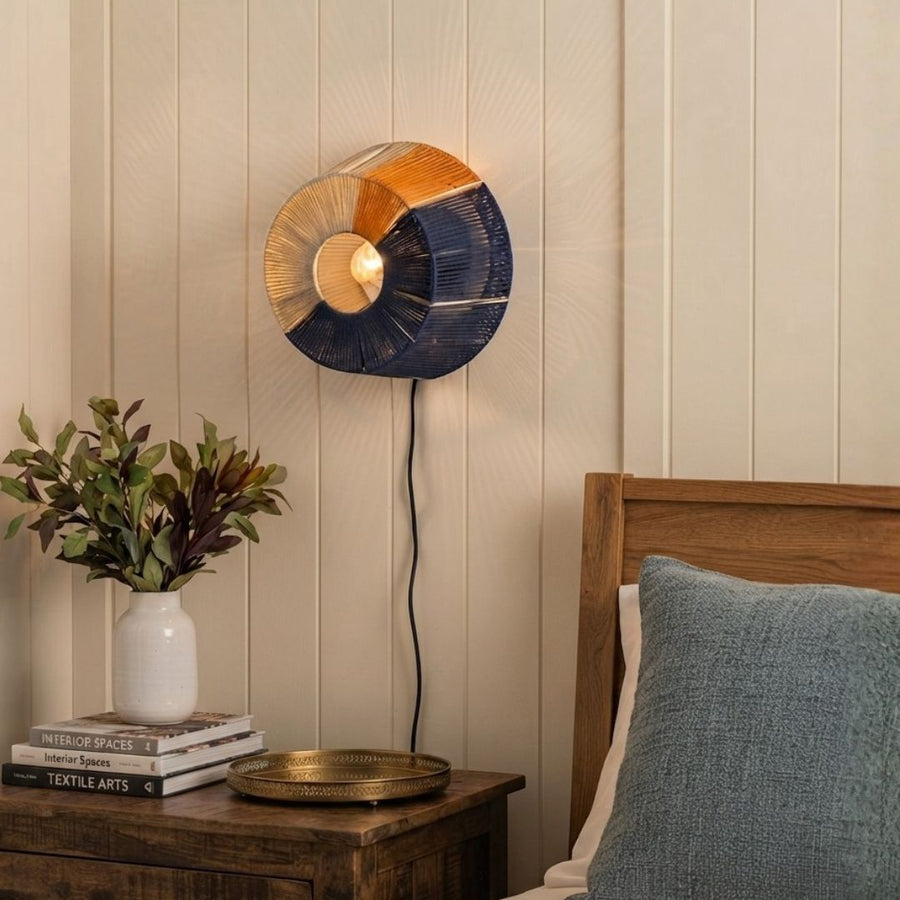 A modern circular wall lamp with a split orange and navy blue shade, mounted on a cream paneled wall above a wooden bedside table.