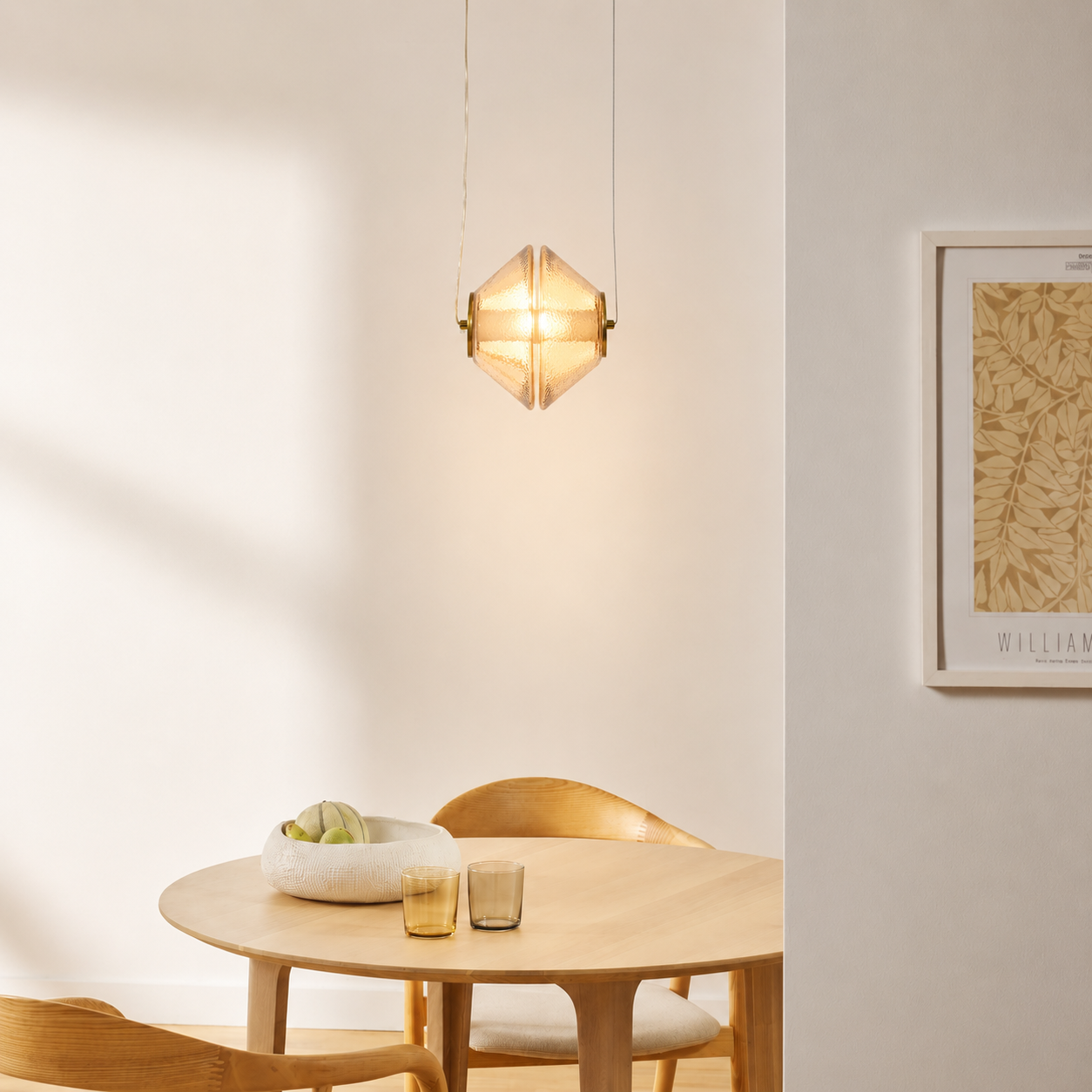 A modern geometric pendant light with a warm glowing glass shade hangs above a round wooden dining table in a minimalist room with cream walls and light wood chairs.