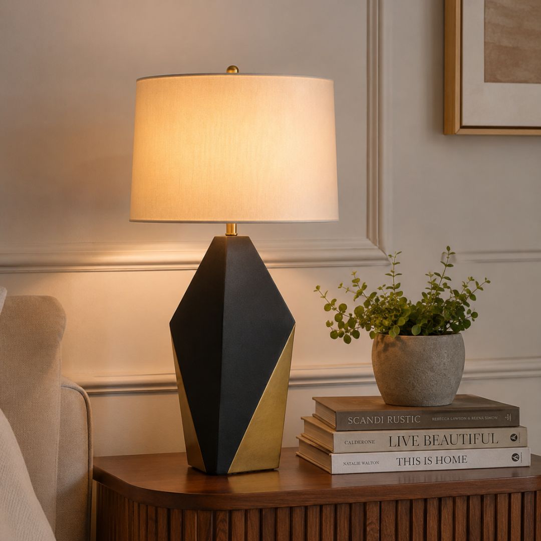 A modern table lamp with a geometric black and gold base and a warm white drum shade, styled on a wooden side table next to a potted plant and books in a cozy room setting.
