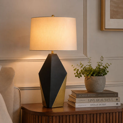 A modern table lamp with a geometric black and gold base and a warm white drum shade, styled on a wooden side table next to a potted plant and books in a cozy room setting.