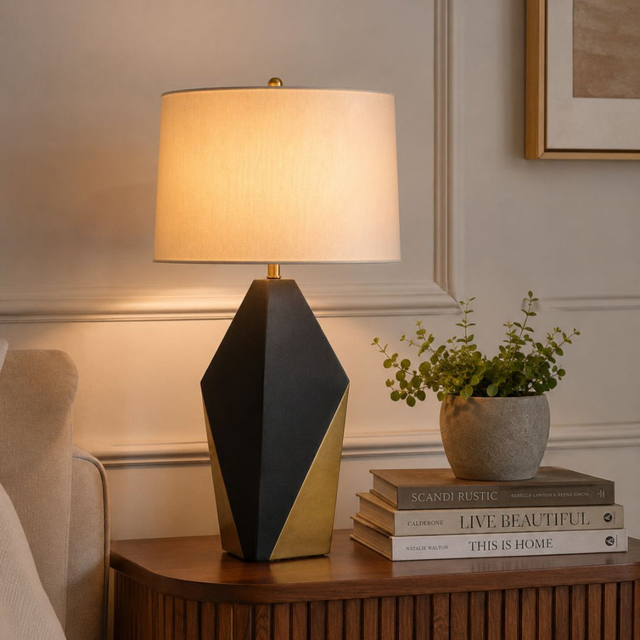 A modern table lamp with a geometric black and gold base and a warm white drum shade, styled on a wooden side table next to a potted plant and books in a cozy room setting.