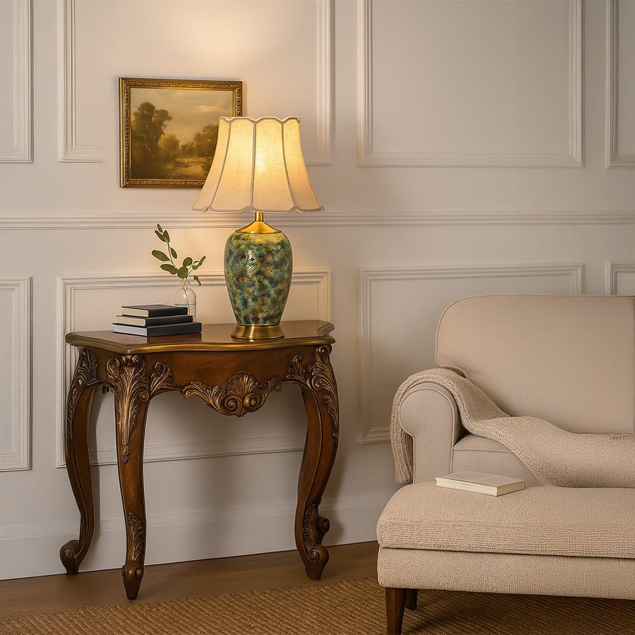 Elegant ceramic table lamp with green and gold artistic base shown with light on placed on a carved wooden console creating a warm classic living room look.