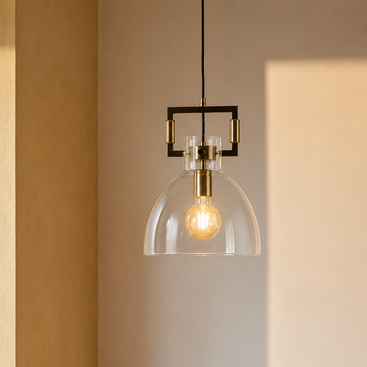 A modern pendant light with a clear glass dome shade, a brass-finished metal frame, and a visible warm Edison bulb, hanging against a beige wall.