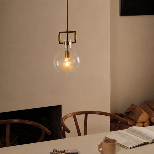 A modern pendant light with a clear glass globe shade and brass hardware hangs over a dining table in a warm, cozy room setting.