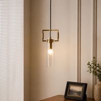 A modern brass pendant light with a clear glass cylinder shade hanging in a warm, lit interior setting next to a window and a small framed picture.