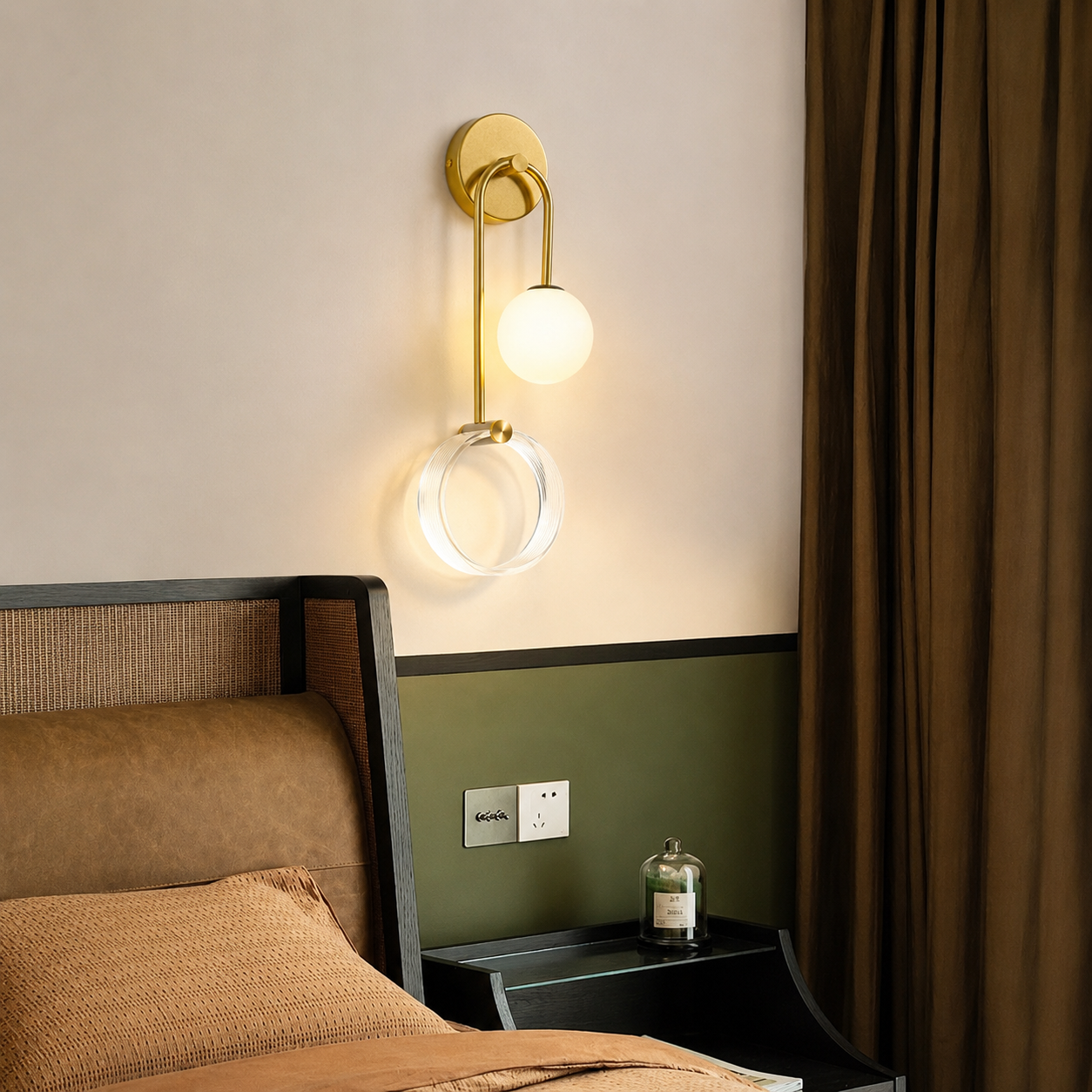 A modern wall-mounted sconce with a gold-finished metal frame, a spherical frosted glass globe, and a clear circular ring, shown lit in a bedroom setting above a bed.