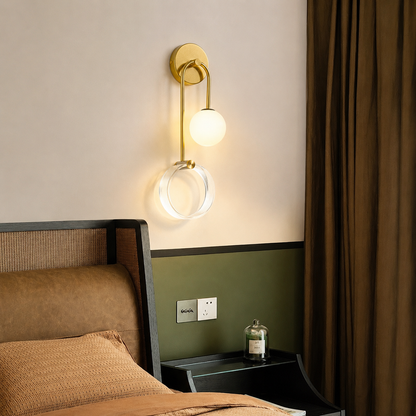 A modern wall-mounted sconce with a gold-finished metal frame, a spherical frosted glass globe, and a clear circular ring, shown lit in a bedroom setting above a bed.