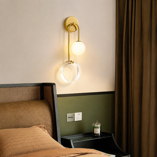 A modern wall-mounted sconce with a gold-finished metal frame, a spherical frosted glass globe, and a clear circular ring, shown lit in a bedroom setting above a bed.