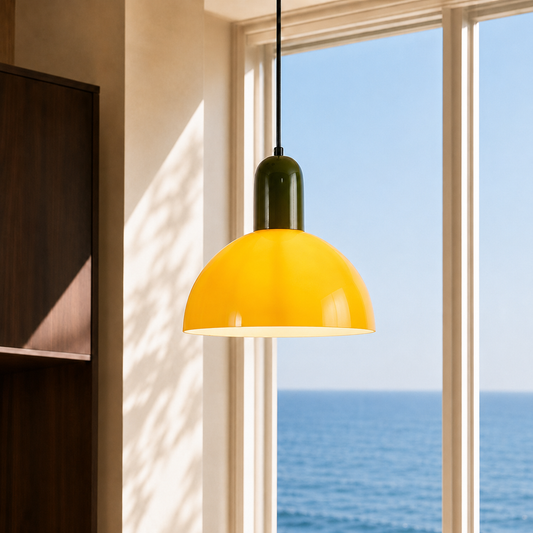 A modern pendant light with a glossy yellow dome shade and a dark olive green neck, hanging in front of a large window with a view of the blue ocean.