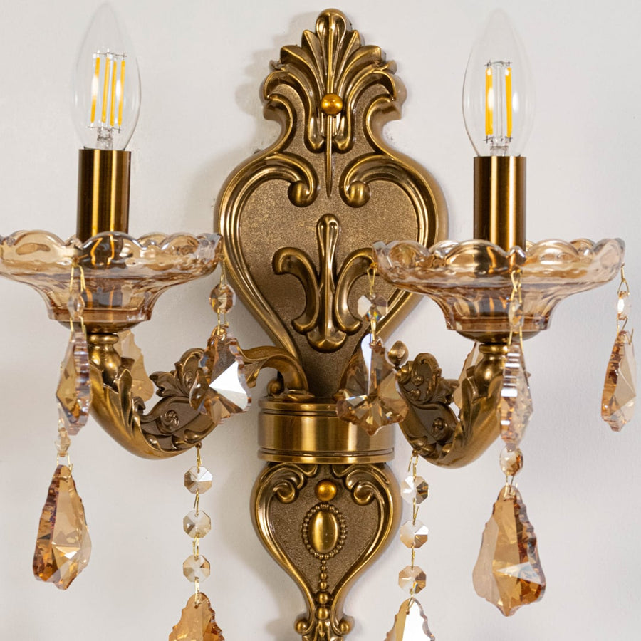 Closeup of gold 2 light wall sconce with crystal drops and dual candle style bulbs for classic home interiors.