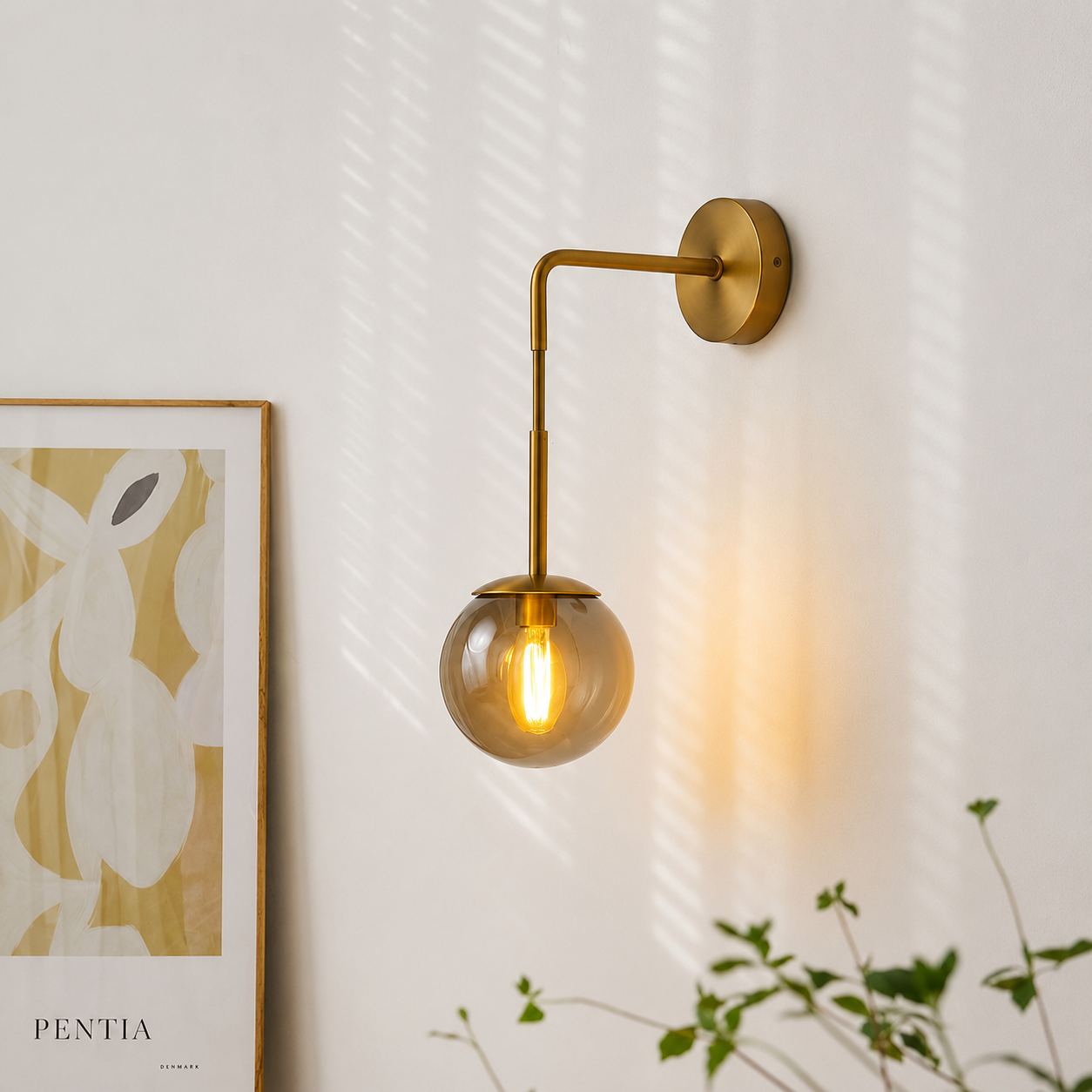A modern wall-mounted sconce with a round glass globe shade and a warm glowing bulb, attached to a white wall with soft window-light shadows. Part of a framed abstract print and green plant leaves are visible at the edges.