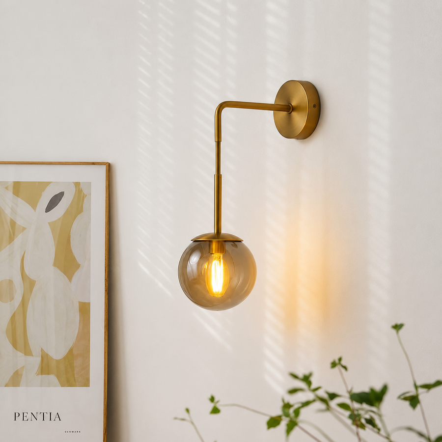 A modern wall-mounted sconce with a round glass globe shade and a warm glowing bulb, attached to a white wall with soft window-light shadows. Part of a framed abstract print and green plant leaves are visible at the edges.