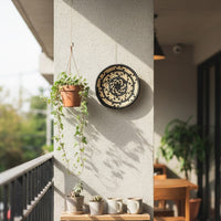 Decorative circular wall art with nature motif placed near window enhancing indoor greenery setting with warm tones and elegant handcrafted design appeal.