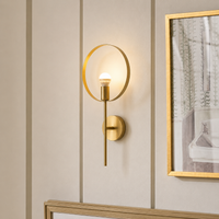 A modern wall sconce light fixture with a gold finish, featuring a circular ring shade and a vertical bar with an illuminated bulb, mounted on a beige paneled wall next to a framed print.