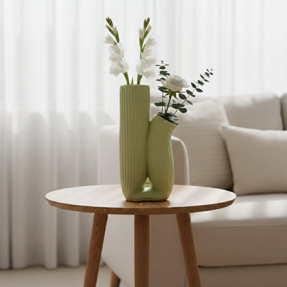 Light green ceramic ribbed dual vase placed on round side table with flowers styled in bright living room.