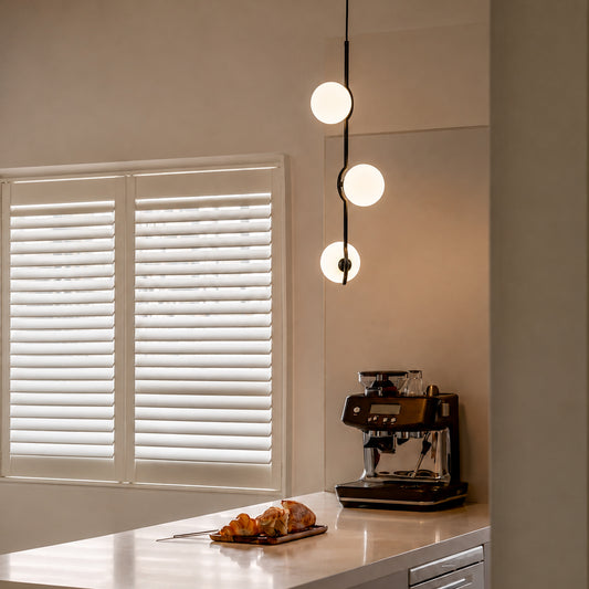 A modern three-light linear pendant lamp with glowing white spherical bulbs hangs over a kitchen counter with a coffee machine and croissants, set against a neutral beige wall and white shutters.