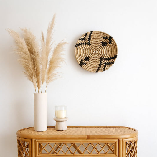 Lifestyle image showing a handwoven round wall basket in natural and black tones displayed on a white wall above a rattan console with pampas grass and soft neutral decor on a white background.