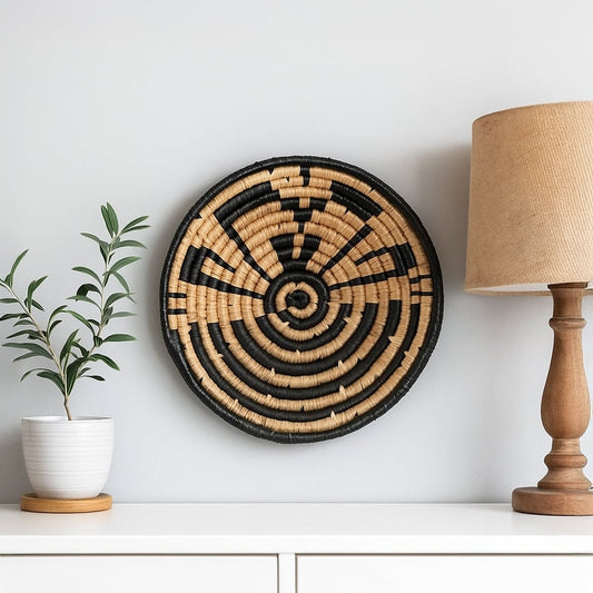 Lifestyle image featuring a handcrafted round woven wall basket in black and natural tones displayed on a clean white wall styled with a table lamp and potted plant for modern decor appeal.