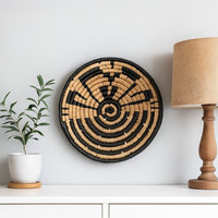 Lifestyle image featuring a handcrafted round woven wall basket in black and natural tones displayed on a clean white wall styled with a table lamp and potted plant for modern decor appeal.
