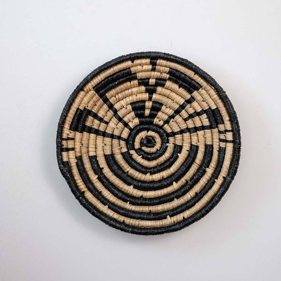 Handwoven seagrass round wall basket decor in natural beige and black with geometric spiral woven pattern on white background.