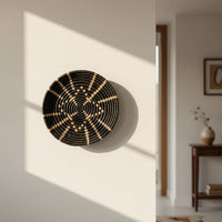 Lifestyle image showing a handcrafted round woven wall basket in black and natural tones mounted on a sunlit neutral wall adding warmth texture and artisanal character to a modern home interior.