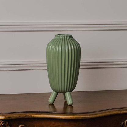Green ribbed ceramic vase standing on a clean white background showcasing its tall form and tripod legs with a smooth matte finish.