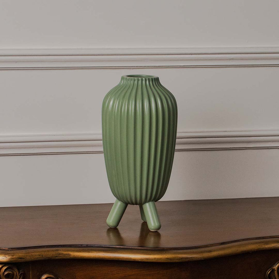 Green ribbed ceramic vase standing on a clean white background showcasing its tall form and tripod legs with a smooth matte finish.