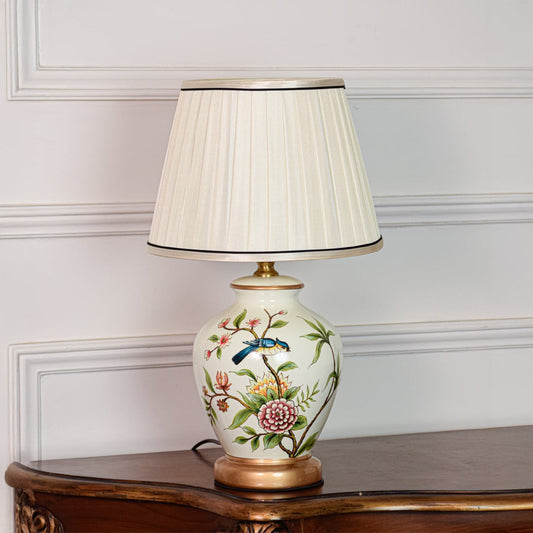 Decorative white ceramic table lamp with floral bird pattern brass base and fabric shade placed on soft off white background.
