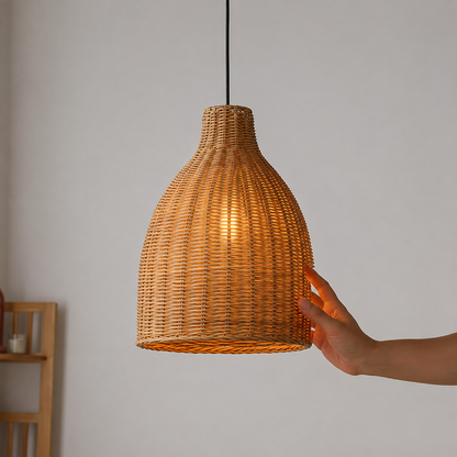 A woven rattan pendant light shade glowing with warm light, shown hanging against a plain wall with a person's hand touching the side of the shade.