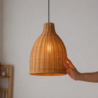 A woven rattan pendant light shade glowing with warm light, shown hanging against a plain wall with a person's hand touching the side of the shade.