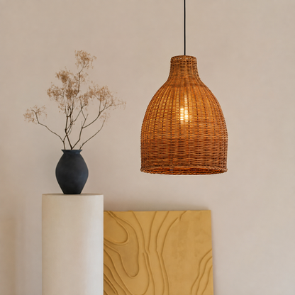 A woven rattan pendant light with a warm glowing bulb inside, hanging in a minimalist room setting next to a white pedestal with a dark blue vase of dried branches and a yellow abstract canvas.