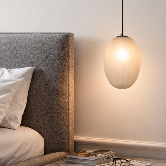A modern oval pendant light with a ribbed frosted glass shade, glowing with a warm light next to a bed with a textured gray headboard.