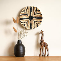 A handwoven natural and black rattan wall decor plate styled on a beige wall above a wooden table with a black vase and giraffe sculpture creating warm modern decor.
