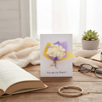 A handmade greeting card featuring a mini yellow and purple dried flower bouquet resting on a rustic wooden study desk.