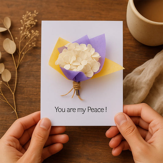 Hands holding a greeting card with ivory paper flower bouquet and message you are my peace on wooden table setup.