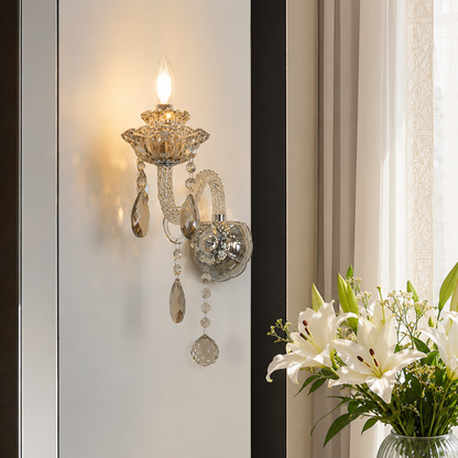 A crystal and gold wall sconce with hanging teardrop beads, shown lit on a white wall next to a vase of white lilies.