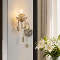 A crystal and gold wall sconce with hanging teardrop beads, shown lit on a white wall next to a vase of white lilies.