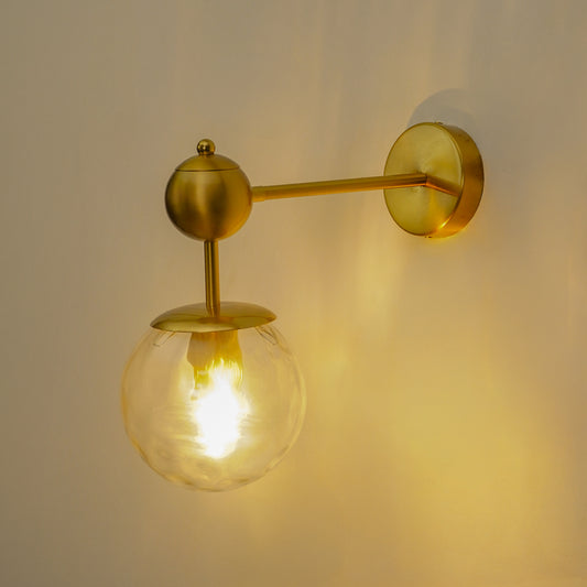 A wall-mounted brass sconce with a spherical glass shade glowing warm yellow, shown against a plain beige wall.